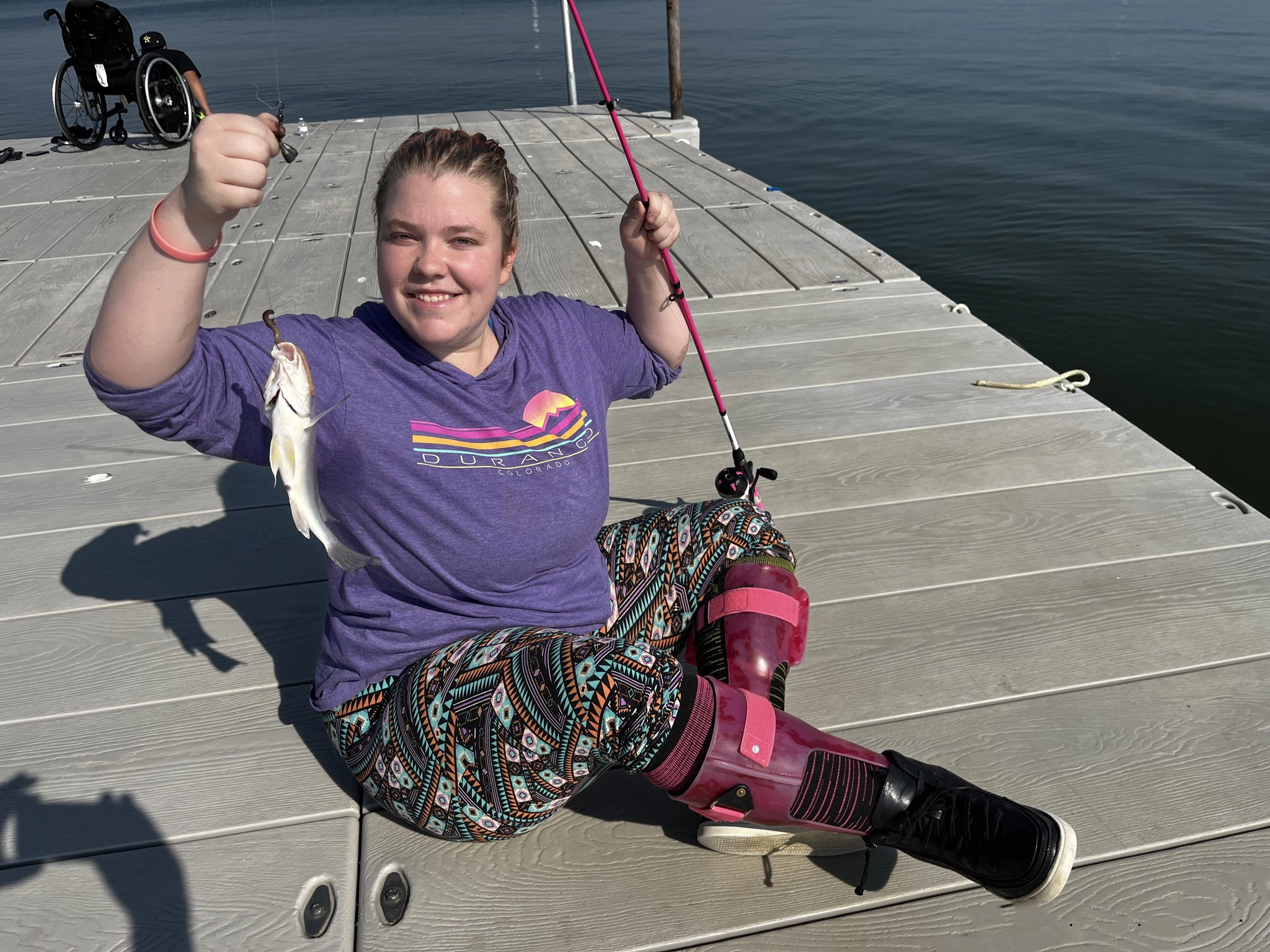 A RISE participant holds up a fish and fishing rod on a dock.