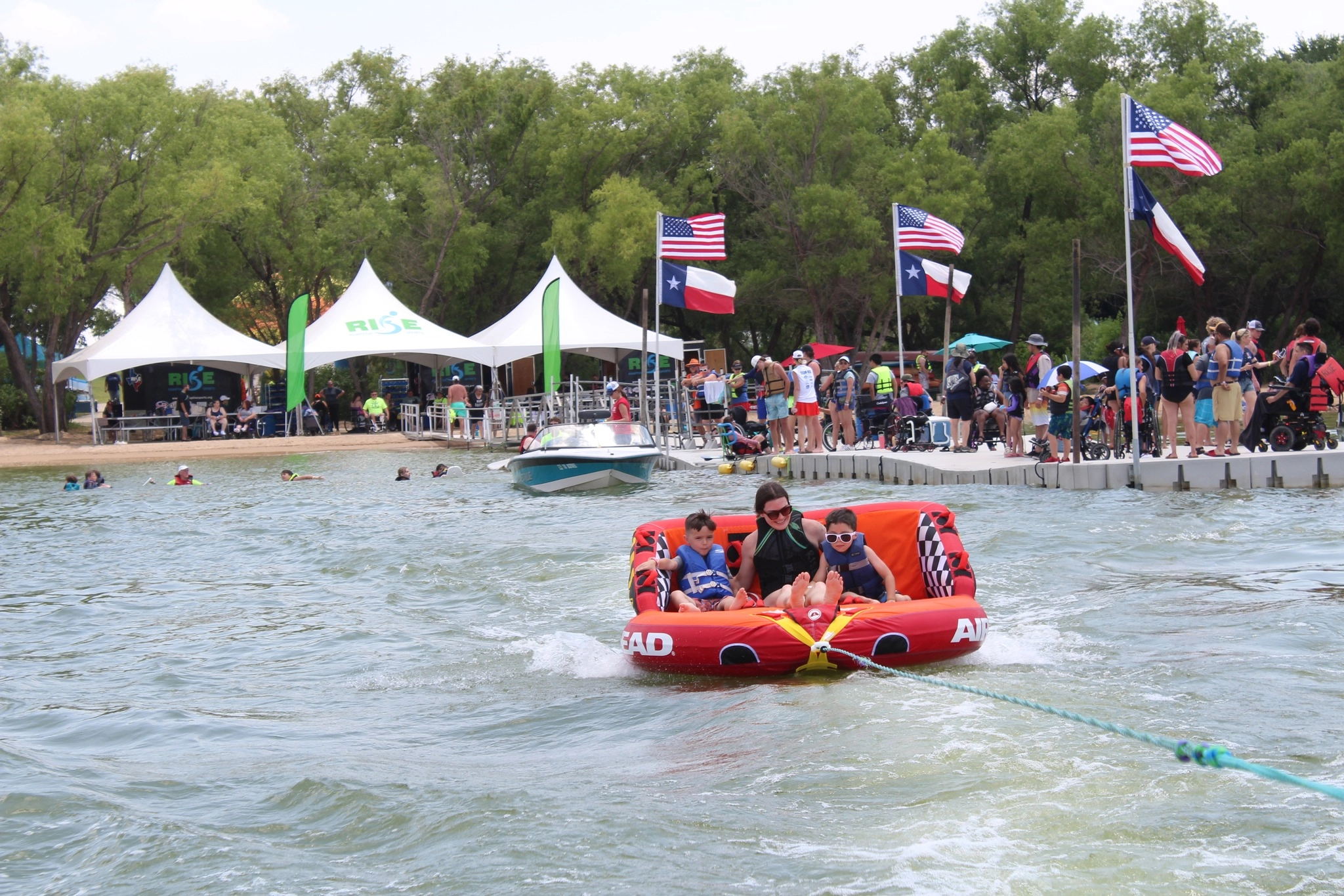 A RISE group gathers at the lake while three people ride a tube in the foreground.
